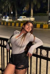 Young woman leaning against a roadside railing at night, wearing a grey top and black shorts, looking to the side under street lights.