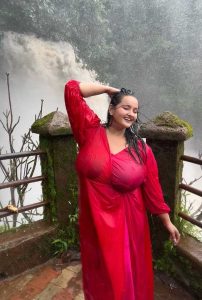 Rimi Busty Escort posing in a flowing red dress by a scenic waterfall, showcasing confident curves and premium escort presence in Siliguri.