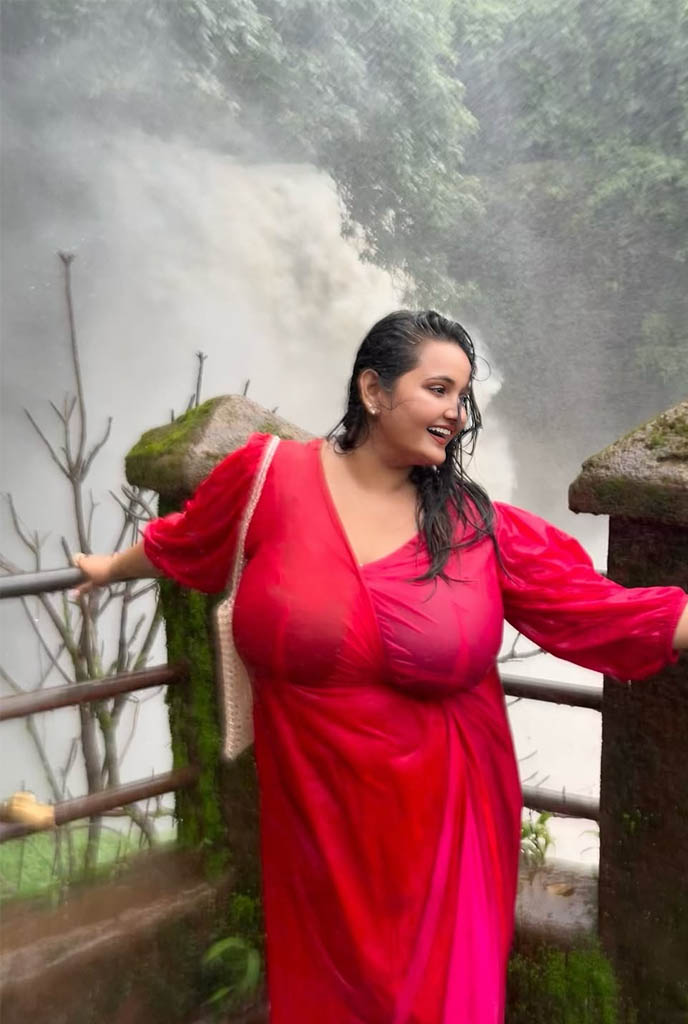 Rimi Busty Escort standing gracefully in a red dress with waterfall backdrop, highlighting bold beauty and luxury escort service in Siliguri.