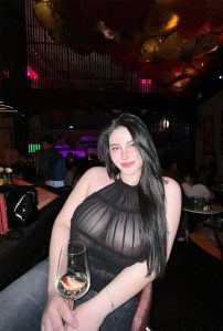 Avnika Sexy Escort enjoying a classy dinner night, wearing a sheer black top and holding a wine glass in a luxury restaurant ambiance.