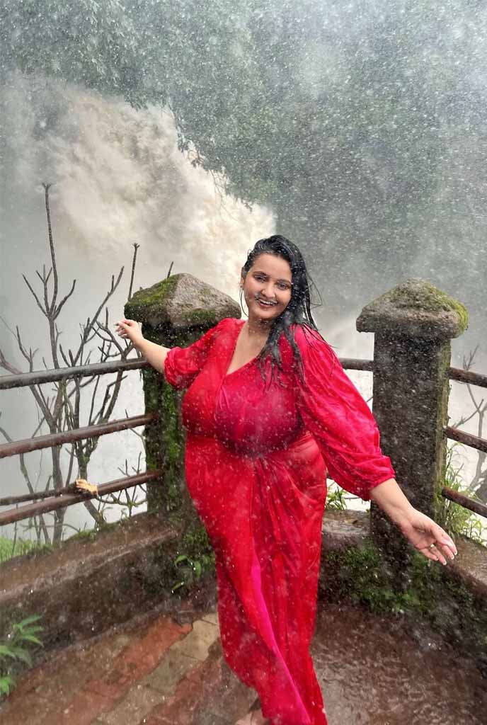 Rimi Busty Escort smiling in a vibrant red dress near a waterfall, reflecting confidence, elegance, and high-profile escort experience in Siliguri.