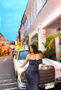 Rimisha Hot Escort posing confidently in a black polka dot bodycon dress while leaning on a vintage white car in a lively evening street setting.